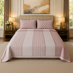 A neatly made bed in a styled bedroom setting, featuring a pink and white patterned quilt with matching pillow shams, flanked by two matching pink and white patterned curtains, gold bedside tables with lamps, and a gold headboard.